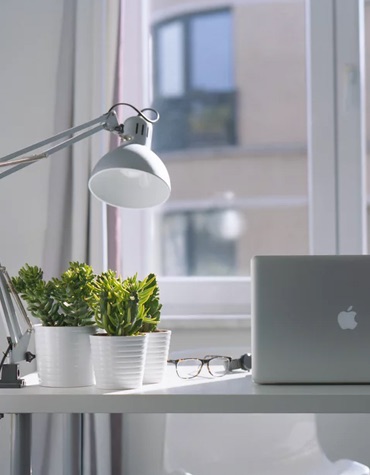 A white desk with plants, a lamp and a macbook in front of a window.