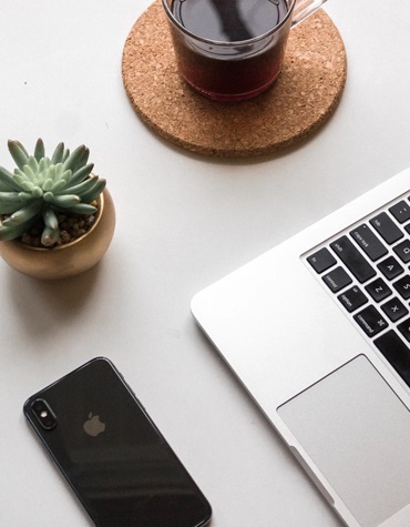 An iPhone, a macbook, a castuc and a cup of tea on a white tabletop.